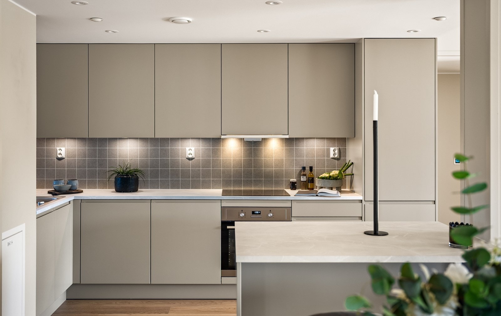 Mellom benkeplate og overskap er det fliser, og overskapene har integrert belysning. Den effektive utformingen gir rikelig med skap- og benkeplass. Praktisk og moderne kjøkkenøy. Galleribilde