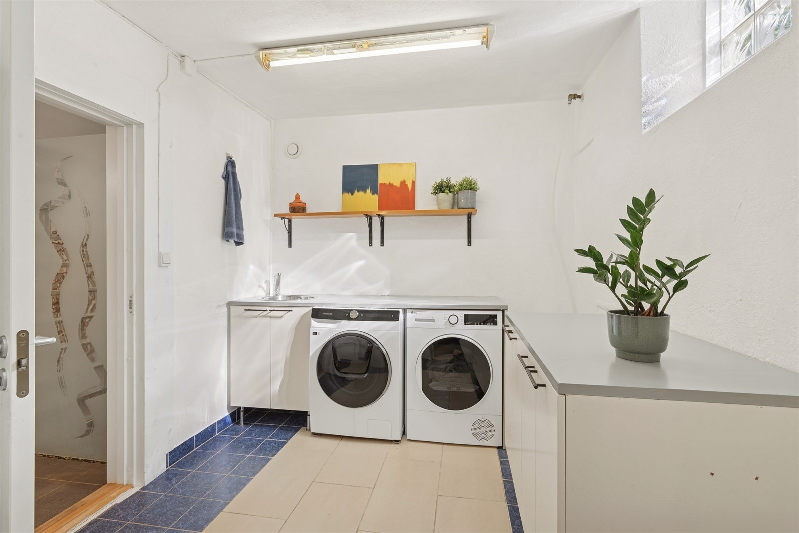 Vaskerom med fliser på gulvet og dør ut til gang/entré. Smart utnyttet med skap og benkeplass, servant og opplegg til vaskemaskin og tørketrommel Galleribilde