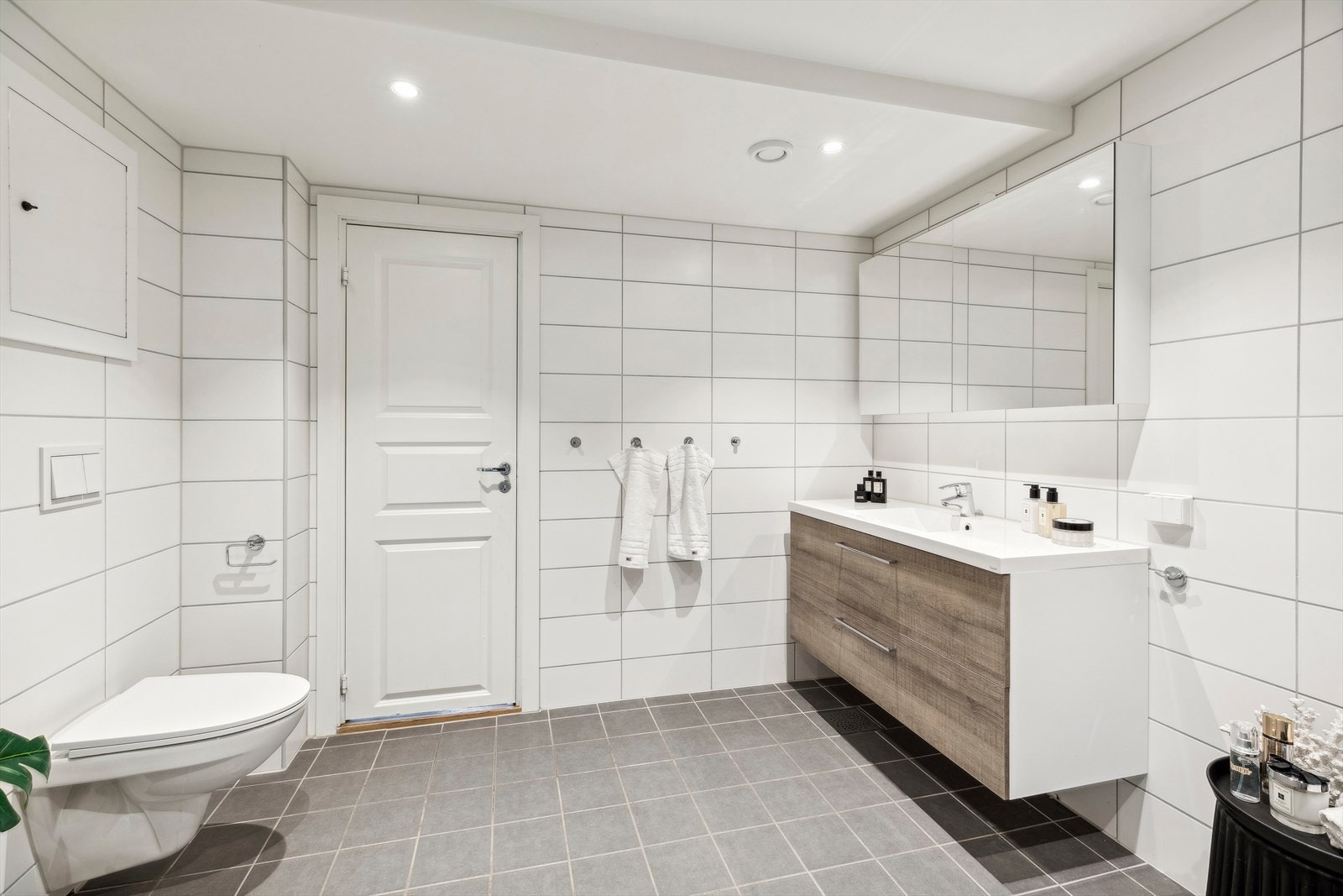 Badet er innredet med veggmontert toalett, dusjhjørne med vegger/dører i herdet glass, servantinnredning og overskap med speilfronter. Galleribilde