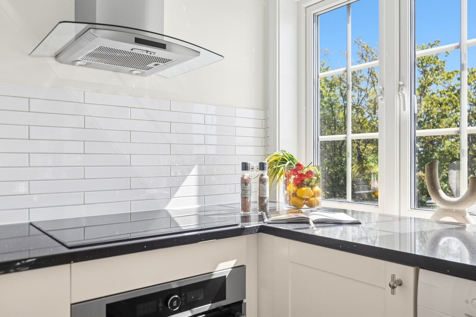 Lekkert kjøkkenhjørne med vindu over benken som gir utsyn mot hagen og rikelig med naturlig lys. Fliser, steinbenk og ventilator setter en moderne ramme. Galleribilde