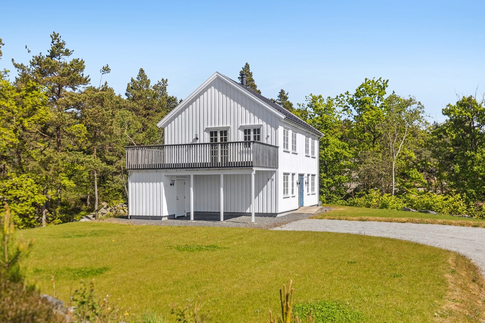 Boligen sett fra innkjøringen med gruset gårdsplass, carport og overbygd inngangsparti. Eneboligen ligger lunt og usjenert til, omgitt av skog og natur. Galleribilde