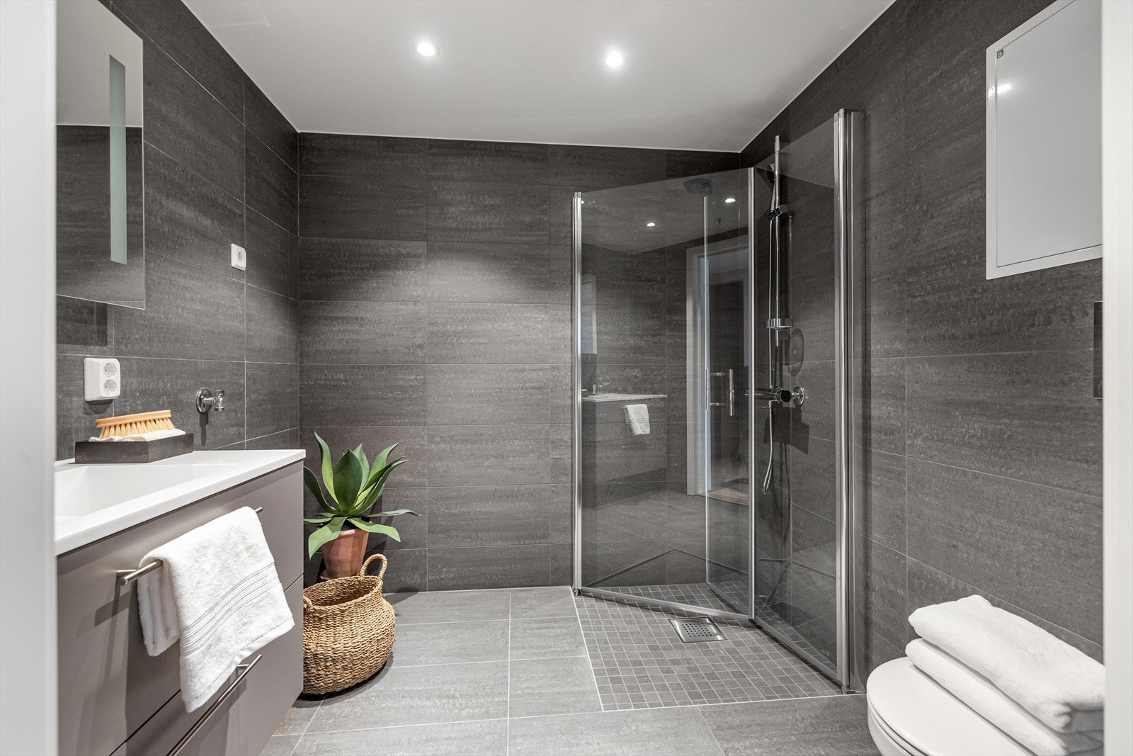 Romslig, flislagt bad fra Pro bad med moderne flisvalg og inredning. Glassdører i dusjen, vegghengt toalett og opplegg for vaskemasking og tørketrommel. Galleribilde