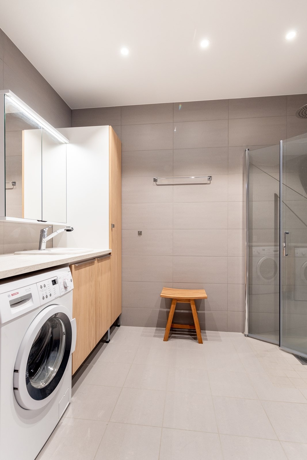 Badet i 1. etasje er helfliset med varmekabler i gulvet, og kombineres som vaskerom. Her er det en romslig baderomsinnredning hvor benkeplaten er praktisk forlenget slik at vaskemaskinen får plass under. Galleribilde