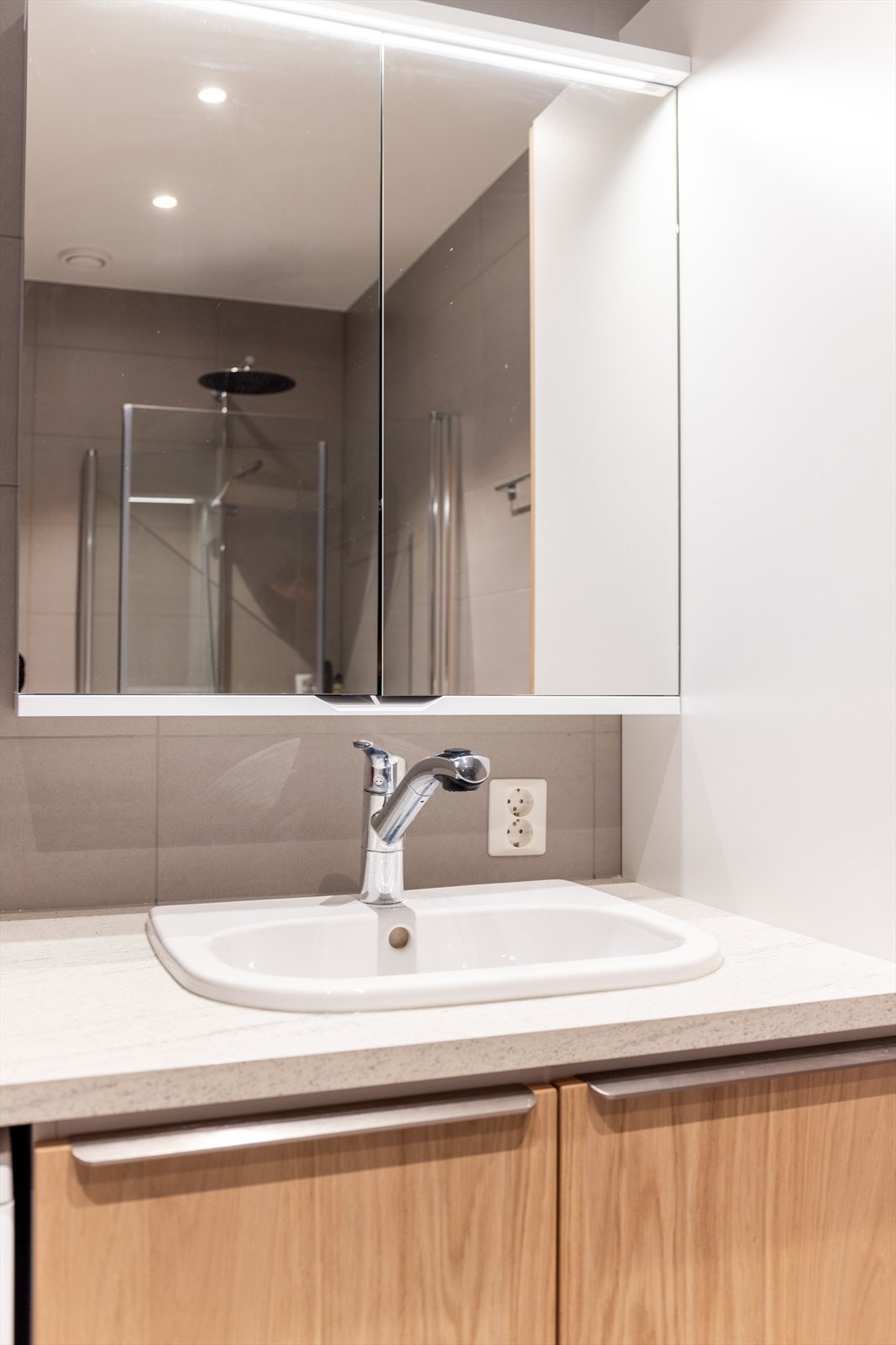 Speilskap med overlys på vegg. Badet er også innredet med dusjnisje med plassbesparende, innfellbare dusjvegger samt veggmontert toalett. Down-light belysning i tak. Galleribilde