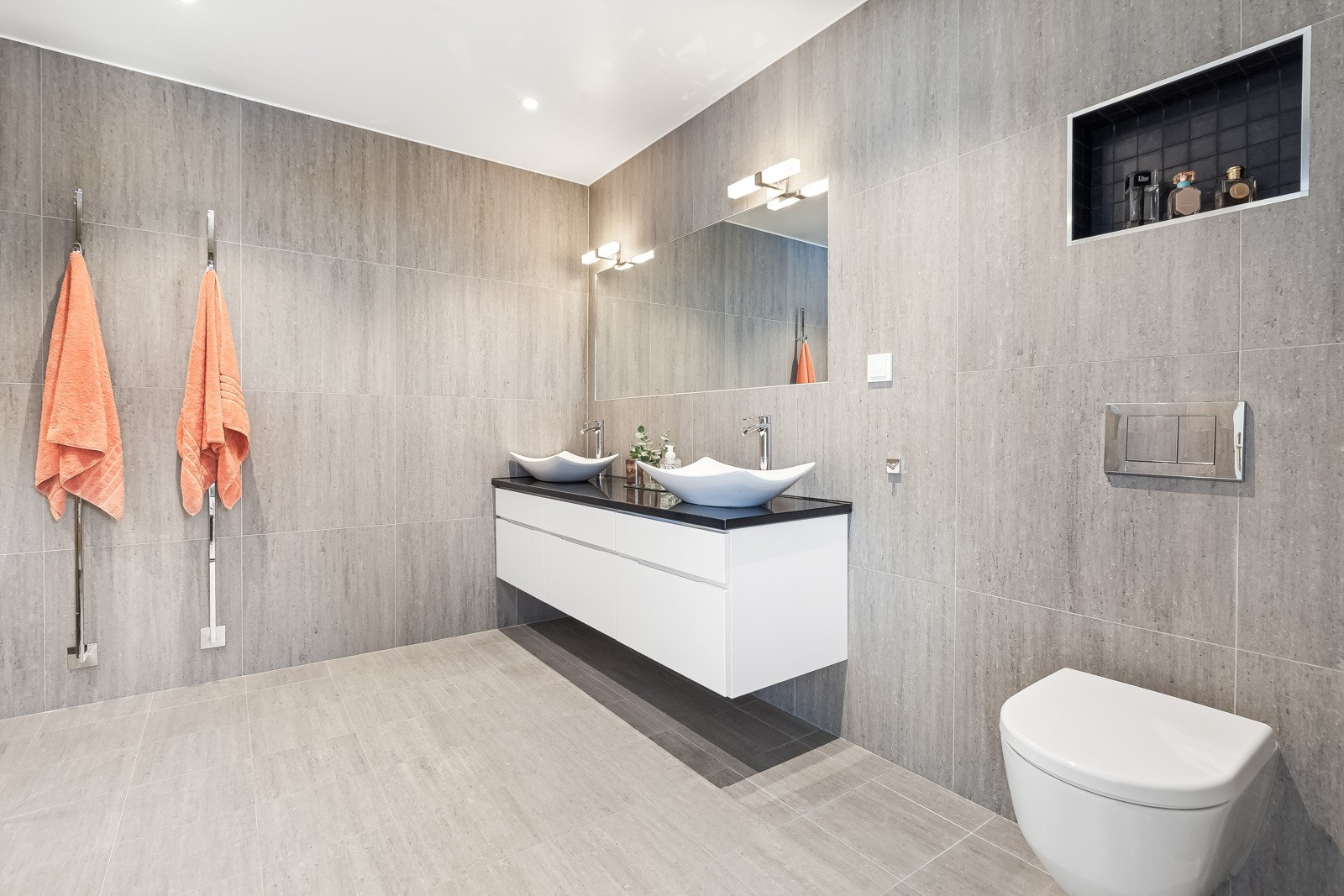 Moderne innredning med dobbel servant og vegghengt toalett. Det er elektriske håndkletørkere på veggen. Granitt benkeplate Galleribilde