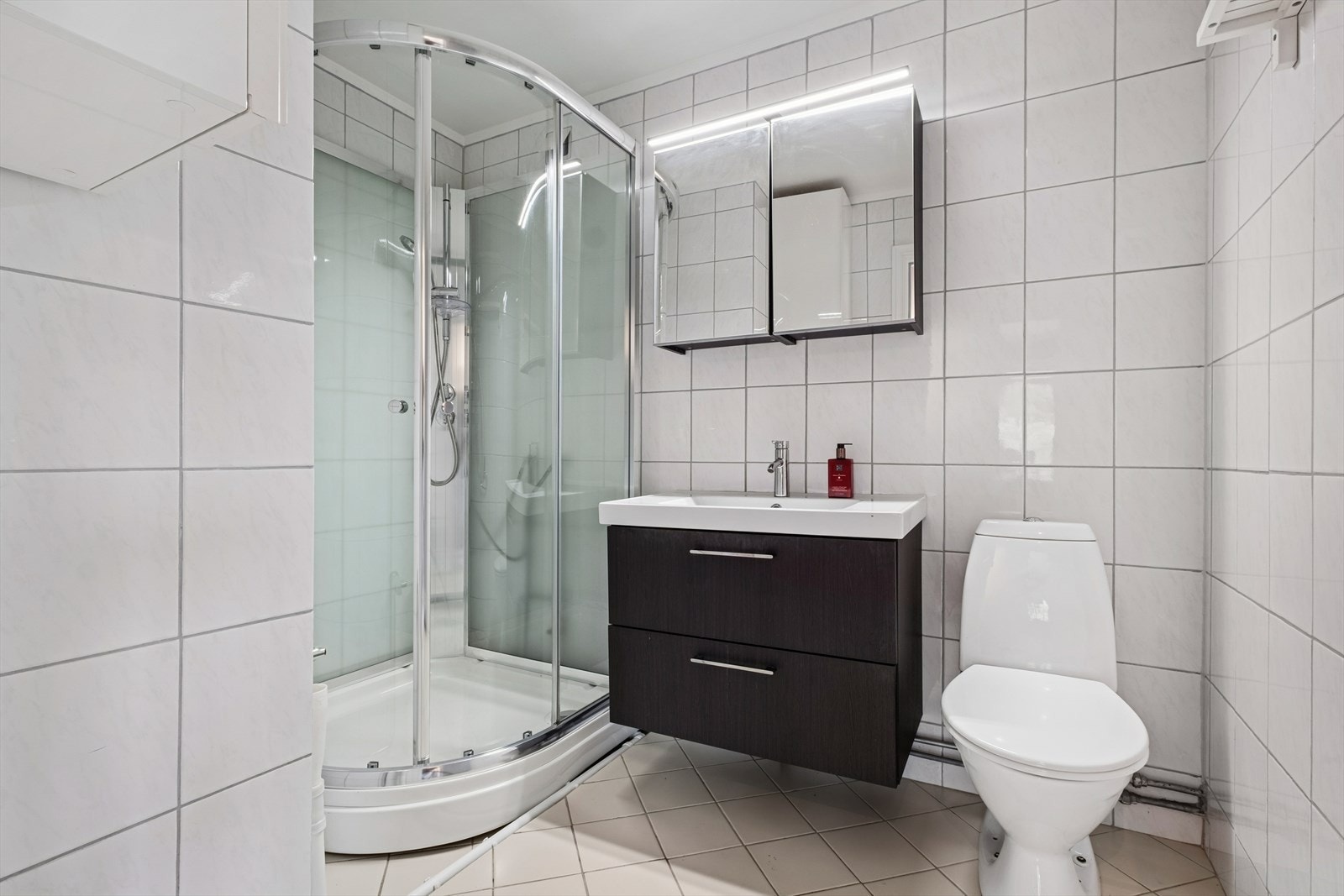 Badet er innredet med fliser på vegger og gulv, klosett, servant nedfelt i innredning, speilskap med lys og dusjkabinett. Galleribilde