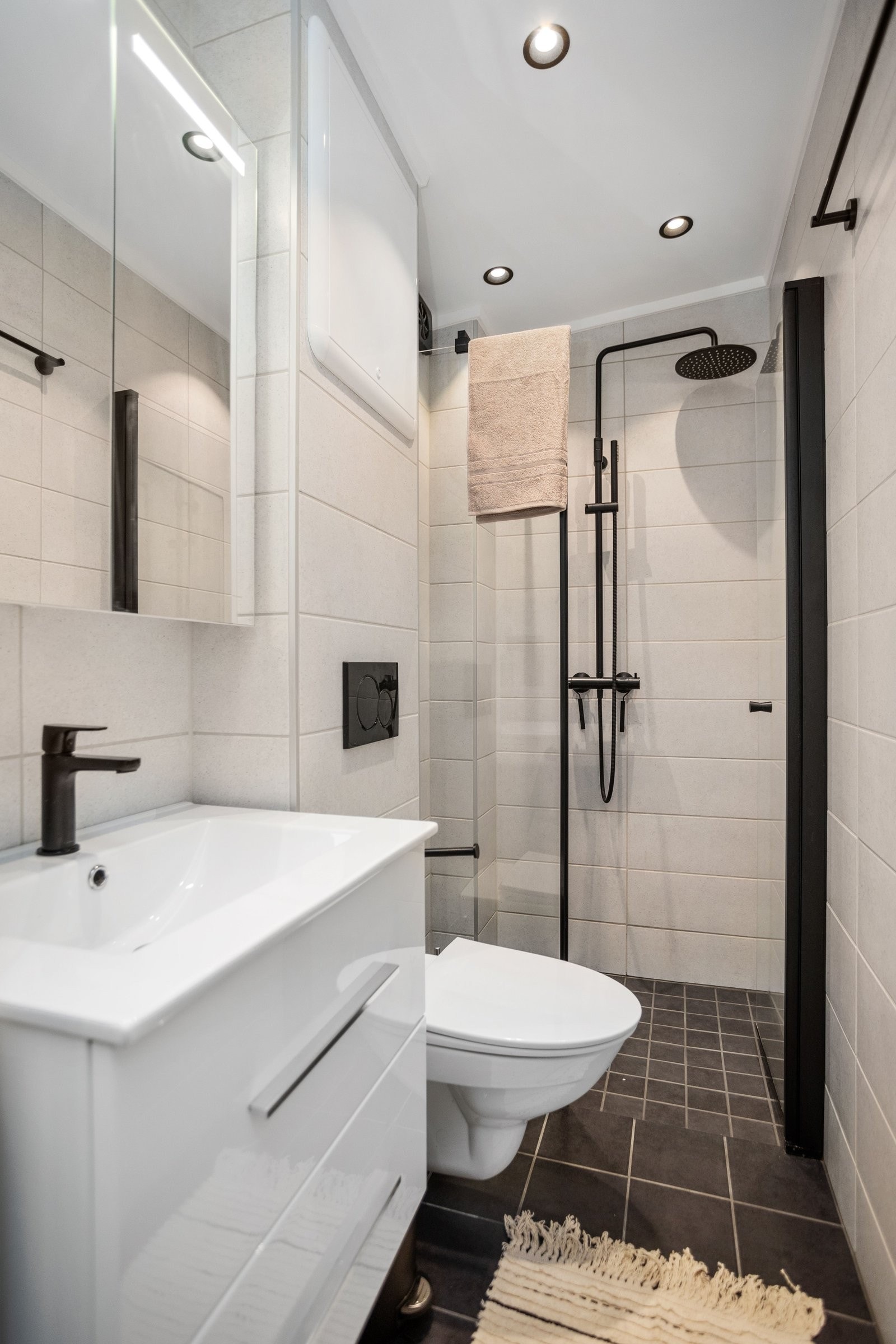 Badet har en stilren utførelse med heldekkende servant på hvit skuffeseksjon, speilskap med integrert lys og vegghengt toalett. Dusjsonen byr på både regnfalls- og hånddusj bak elegante glassdører – en liten hverdagsluksus. Galleribilde