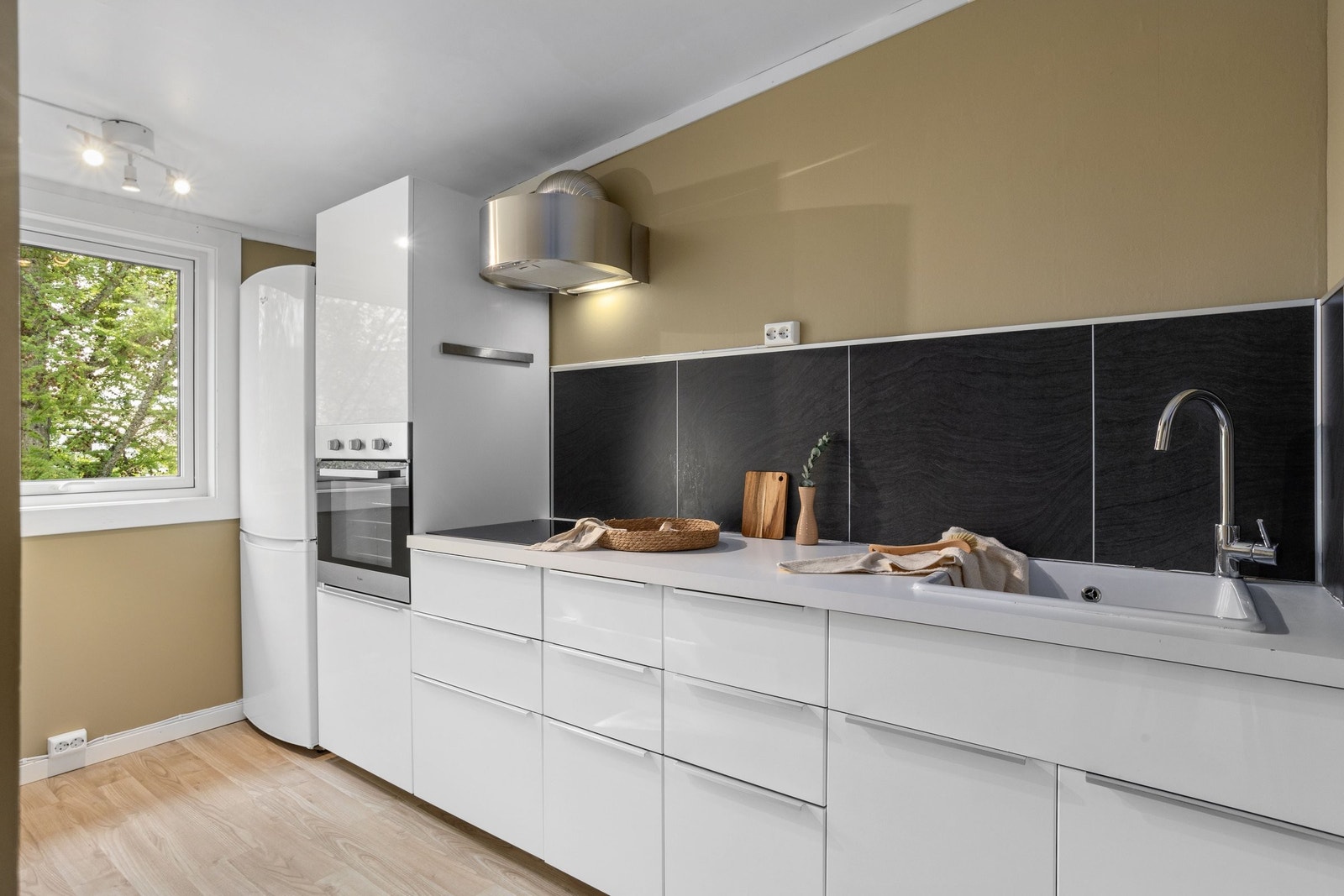 Kjøkkenet har en stilren og moderne utførelse med glatte, hvite fronter, og sorte detaljer. Den effektive utformingen gir godt med arbeids- og oppbevaringsplass, mens det østvendte vinduet slipper inn naturlig morgenlys Galleribilde