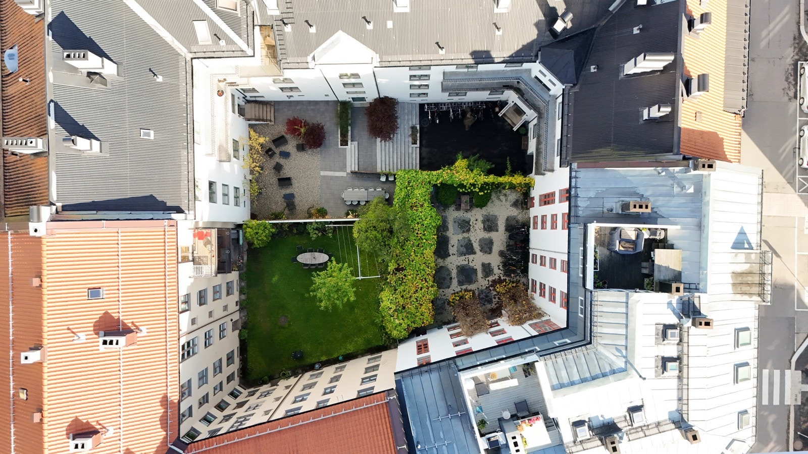 Oversiktsbilde av bygården med et hyggelig, grønt gårdsrom med plen, trær og sitteplasser - et skjermet og innbydende uteområde midt i byen. Galleribilde