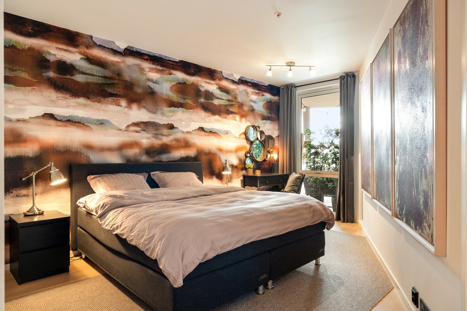 Leiligheten har et romslig soverom holdt i en stilren kombinasjon med lysmalte vegger og lekker tapet mot gyllent parkettgulv, og det er listfrie overganger til lysmalt himling. Det er vindu mot den overbygde balkongen. Galleribilde