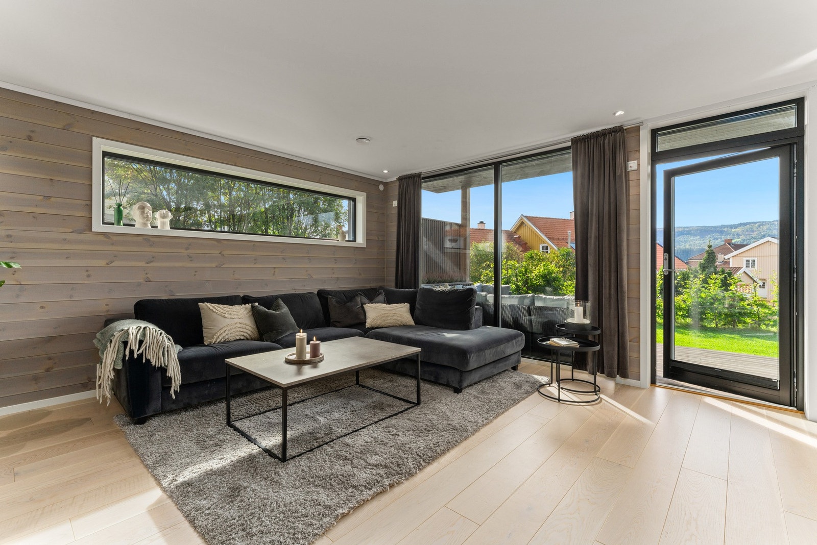 Stuen er romslig, luftig og gjennomstrømmet av dagslys fra store vindusflater som rammer inn utsikten mot grøntarealer og åsene i bakgrunnen. Galleribilde