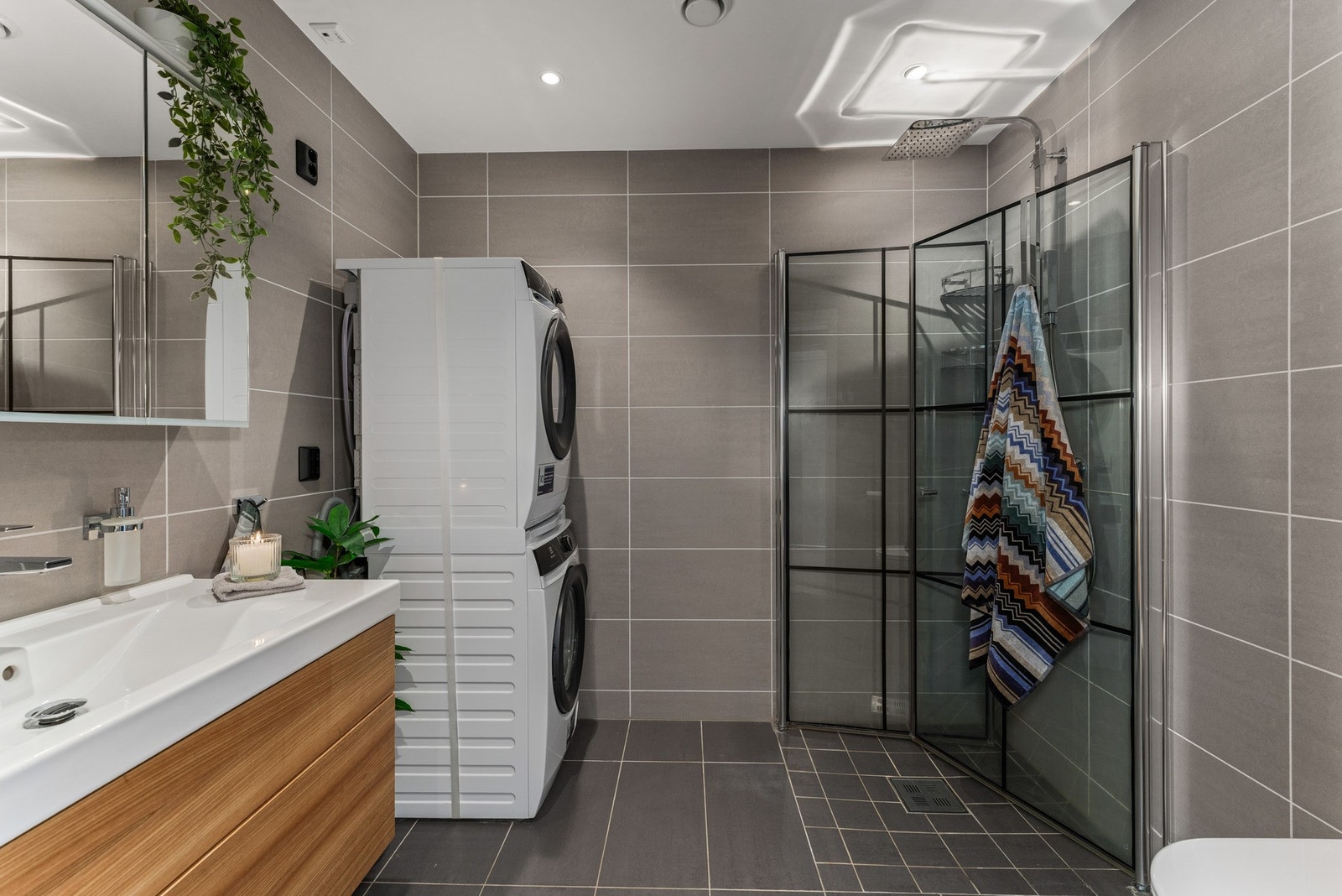 Badet fremstår som lekkert og moderne, med delikate grå fliser, sorte dusjprofiler, vegghengt toalett og servantinnredning i treverk som tilfører varme. Galleribilde