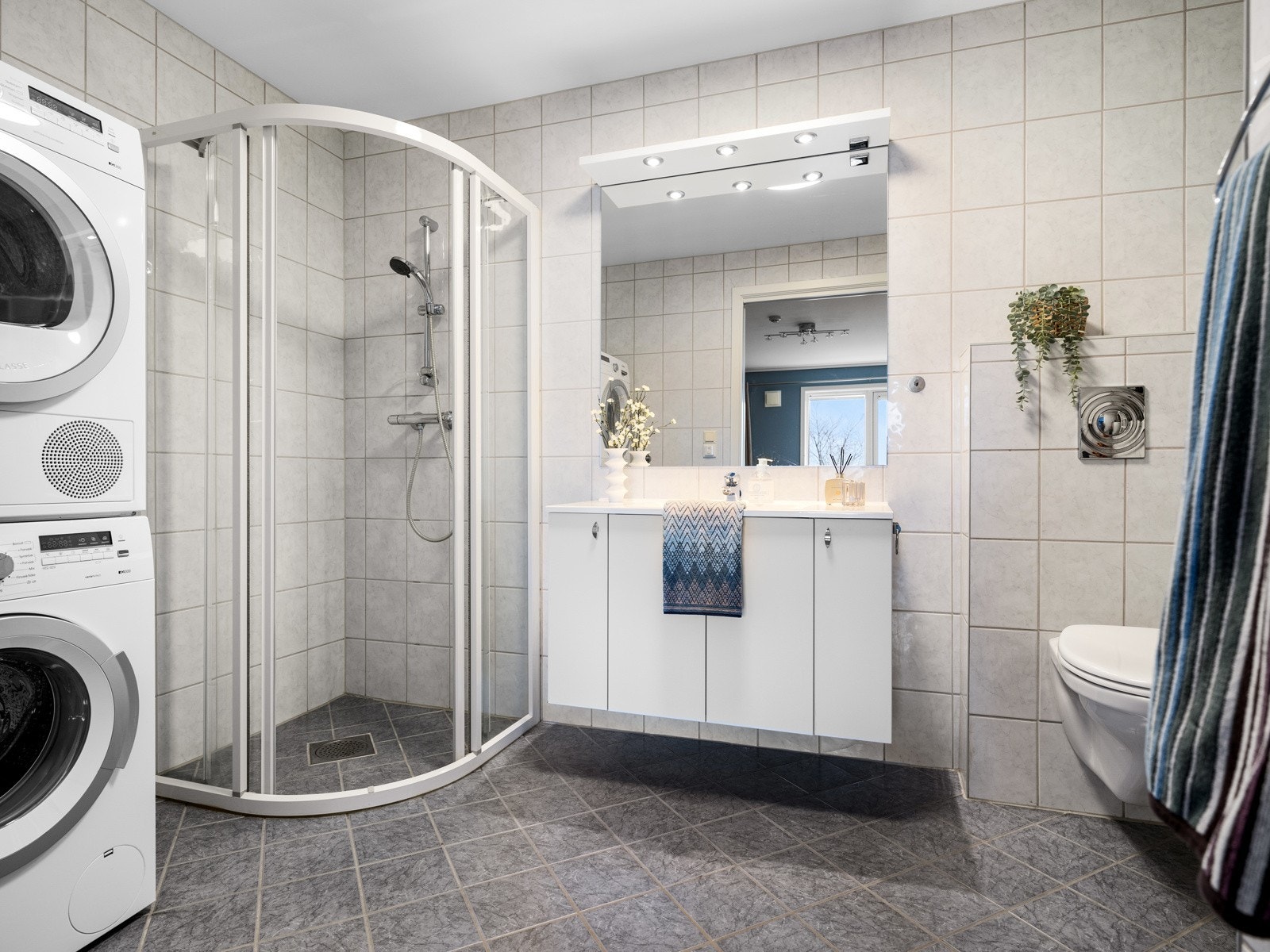 Det andre badet er helfliset i lik utførelse som hovedbadet og har også gulvvarme. Innredningen består av en heldekkende servant på en hvit skapseksjon, speil og overhengende belysning. Badet utstyrt med dusjhjørne, toalett og opplegg til vaskemaskin. Galleribilde