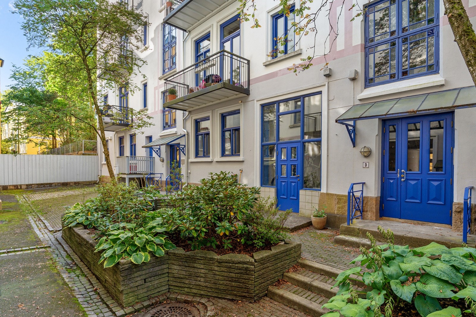 Pen og opparbeidet bakgård belagt med belegningsstein og beplantning i form av planter og trær. I bakgården er det en felles sykkelparkering og vannslange til spyling av sykler eller annet utstyr. Galleribilde