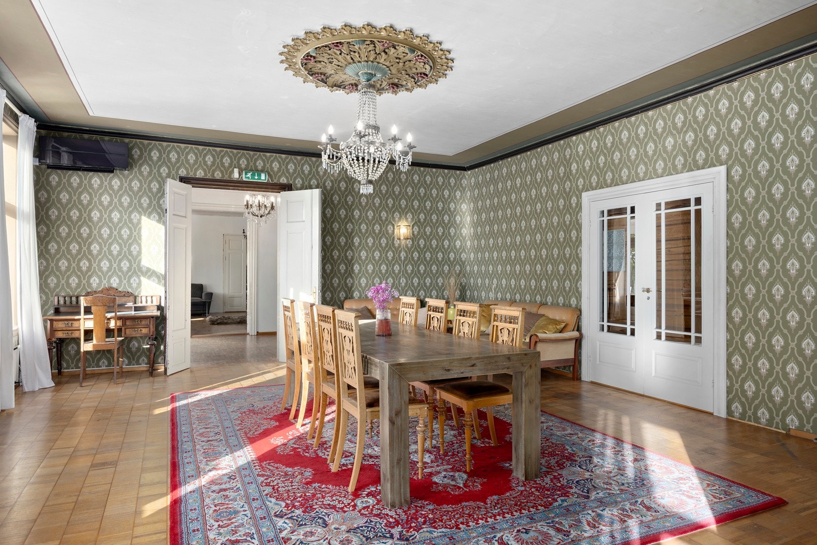 Storstuen fremstår som boligens hjerte, med klassisk grønn tapet, dekorativ takrosett og doble dører inn fra kjøkkenet. Galleribilde