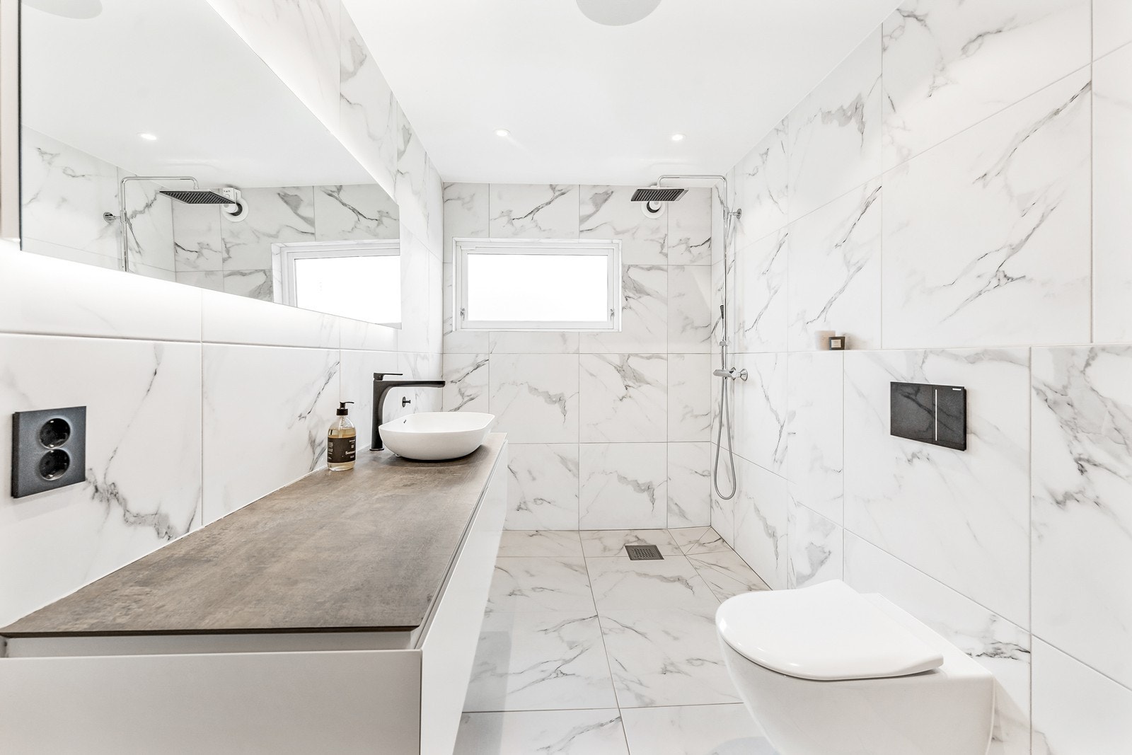 Her får man flislagte vegger, en kontrasterende vegg med eikespiler, dusj uten vegger, vegghengt toalett, samt servant montert på benkeplate med skuffer og speil over. Galleribilde