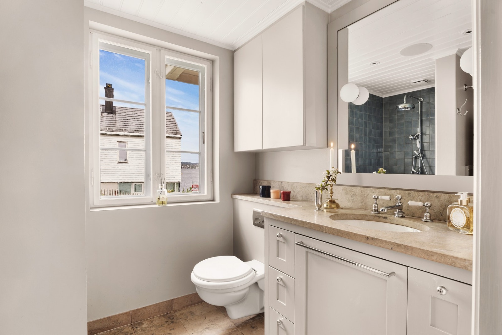 Lyst og delikat baderom med naturstein på benkeplate, klassiske armaturer og vindu som gir både dagslys og glimt av fjorden. Galleribilde