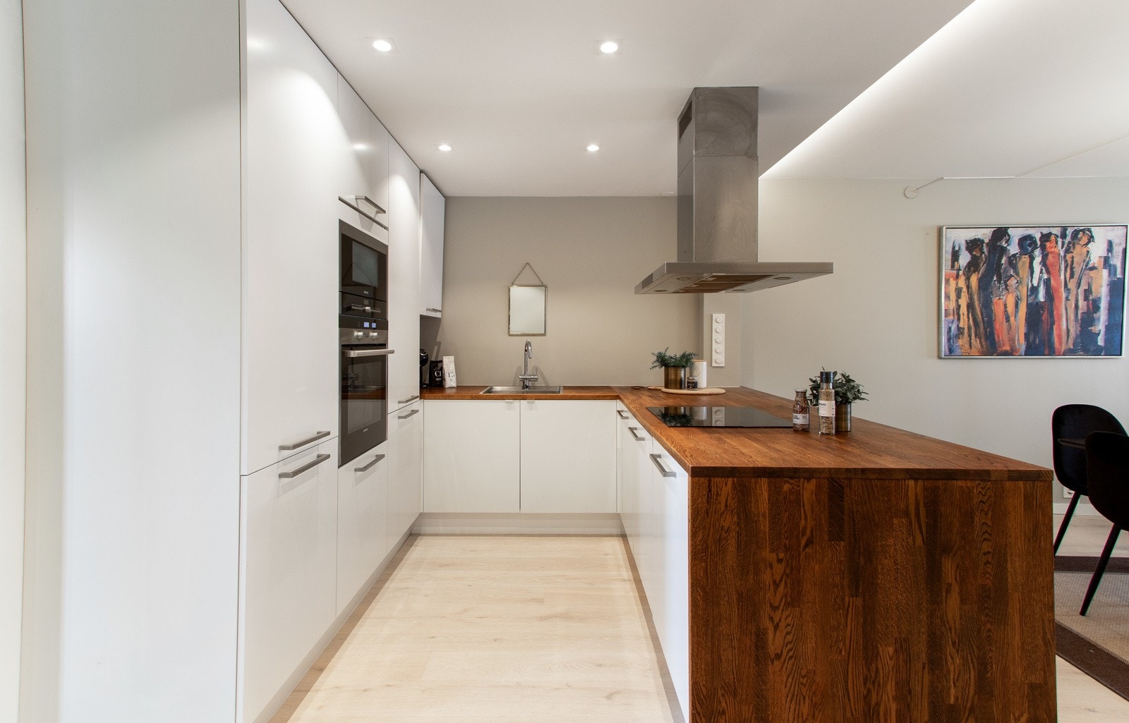 Innredningen har høyskap med hvite, glatte fronter langs innerste vegg, matchende benkeskap, skap ut mot stuen og benkeplate i heltre som er dratt ned langs kanten. Galleribilde