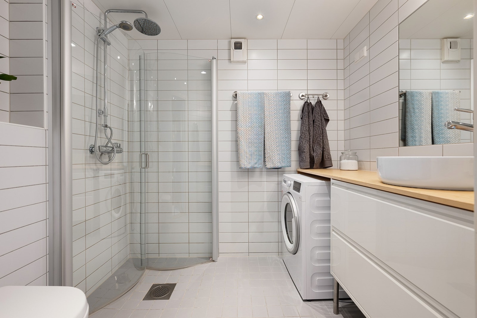 Badet er pent flislagt på gulv og vegger. Badet er innredet med vegghengt klosett, ovenpåliggende servant med servantinnredning, speilskap med overlys og dusjhjørne med dører. Galleribilde