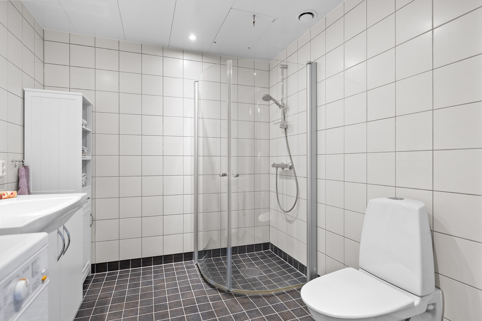 Badet fremstår som et lyst, delikat og praktisk rom med flislagt gulv med gulvvarme som gir en behagelig varme under føttene. Veggene er kledd med moderne fliser, og himlingen har malte metallplater som tilfører rommet et rent og stilfullt uttrykk. Galleribilde