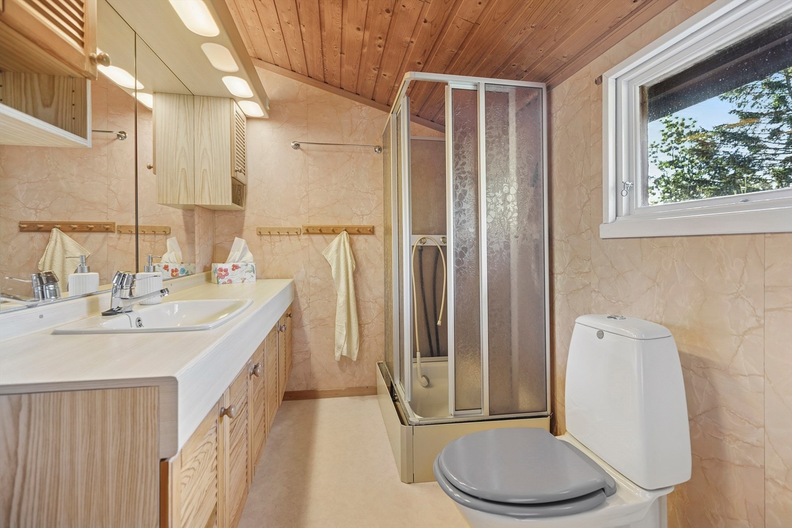 Bad fra 80-tallet med belegg på gulv, våtromsplater på vegger og panel i taket. Badet er utstyrt med toalett, dusjkabinett og baderomsinnredning med servant og speil. Galleribilde