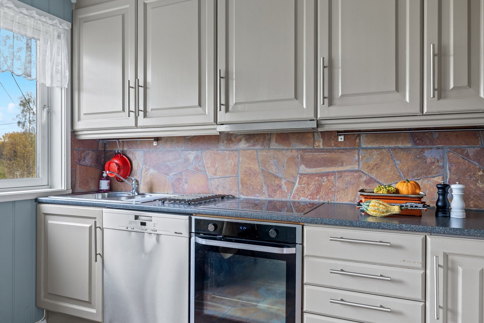 Kjøkkenet er utstyrt med trefiberplater mellom benk og overskap, nedfelt oppvaskkum i rustfritt stål med ett-greps armatur og integrert komfyr med keramisk platetopp. Galleribilde