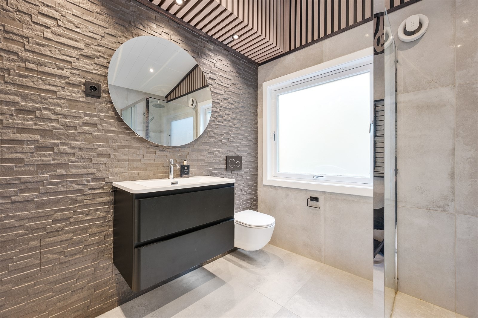 Moderne bad med fliser på gulv og vegger, kontrastvegg som fremhever servant med rundt speil. Praktisk dusjhjørne, vegghengt toalett og opplegg til vaskemaskin for en funksjonell og moderne løsning. Galleribilde