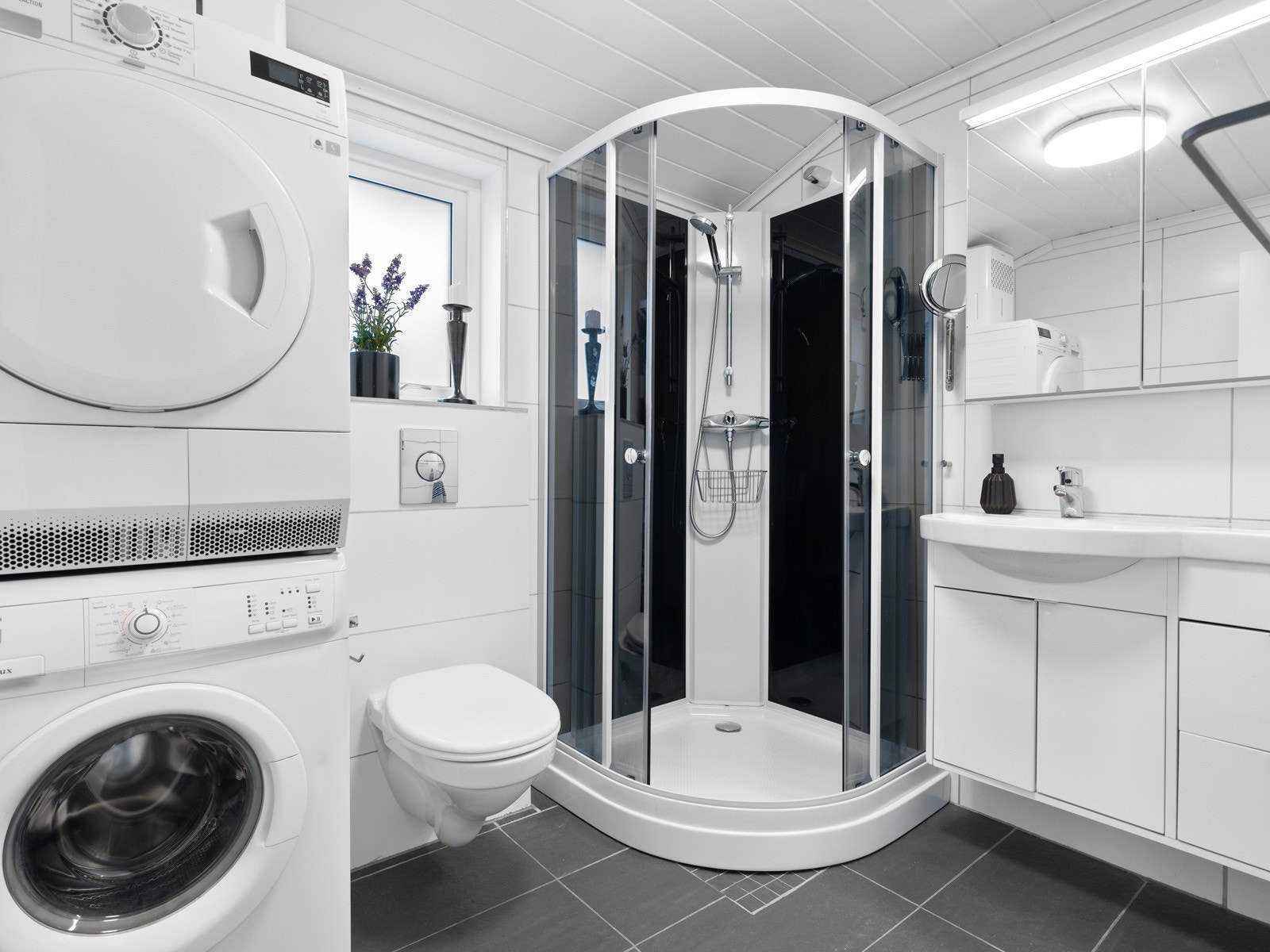 Flislagt bad med plass til vaskemaskin, tørketrommel, vegghengt toalett, dusjkabinett og lys baderomsinnredning. Galleribilde