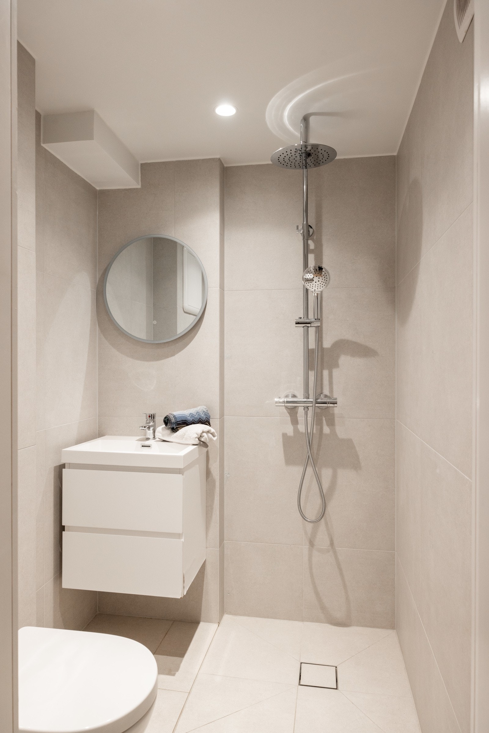 Bad III er flislagt med fliser fra Fagflis. Her finner du servantmøbel med to skuffer, speil med belysning, samt toalett og dusjløsning med både regnfalls- og håndholdt dusj - en komplett og stilren løsning for gjester eller beboere i hybelen