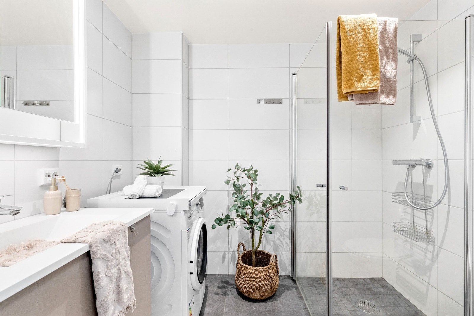 Moderne og flislagt baderom med innfellbare dusjdører, opplegg for vaskemaskin, servant med skap, rørfordelingsskap og veggmontert wc. Galleribilde