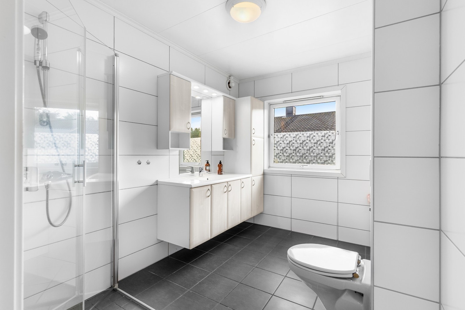 Badet er utstyrt med dusjhjørne med innfellbare dusjdører, servantskap, rørfordelingsskap, veggmontert wc og mekanisk avtrekk. Galleribilde