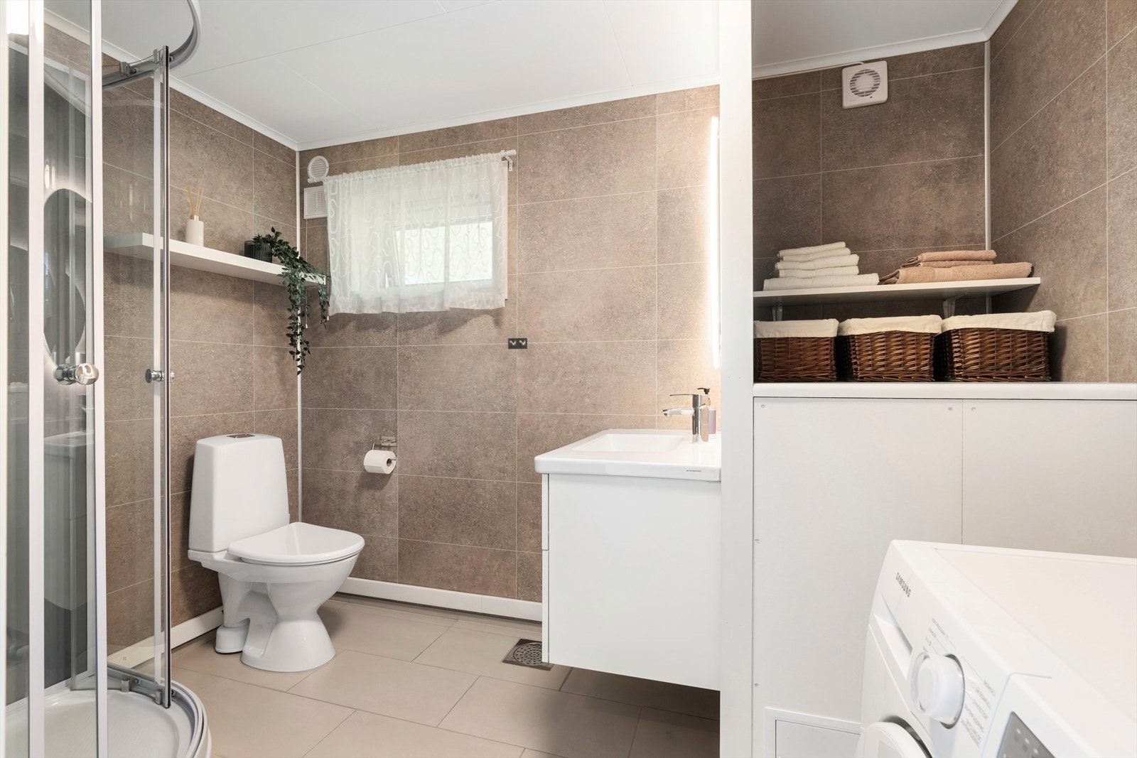 Bad/ vaskerom har flislagt gulv og baderomsplater på vegger, samt dusjkabinett og toalett; elektrisk avtrekksvifte sikrer god ventilasjon. Galleribilde