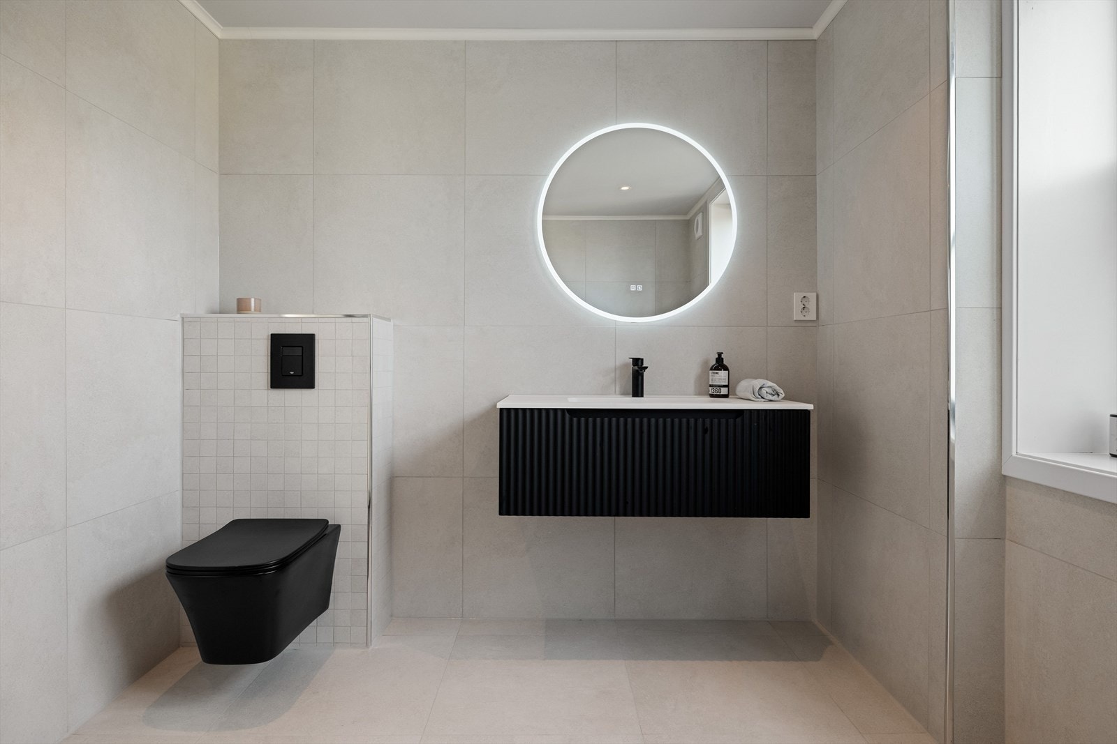 Badet er utstyrt med veggmontert toalett og baderomsinnredning med skuff og heldekkende servant med ett-greps blandebatteri. Badet har en gjennomført stil med mørke detaljer. Galleribilde