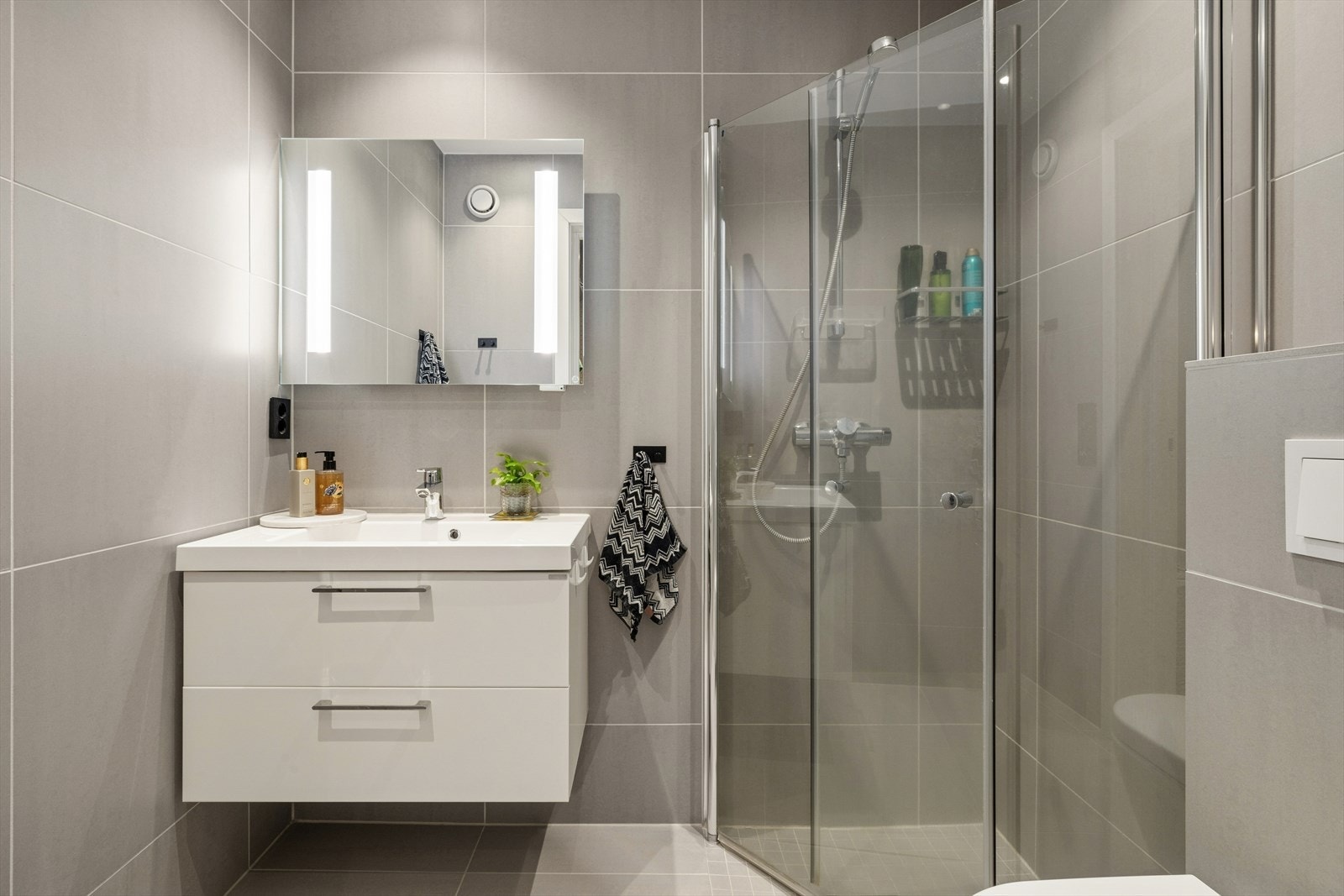 Delikat baderom med tilknytning til omkledningsrom. Utstyrt med servant med skap i glatte fronter, speil og belysning, dusj hjørne med innfellbare dusjdører og vegg montert toalett. Galleribilde