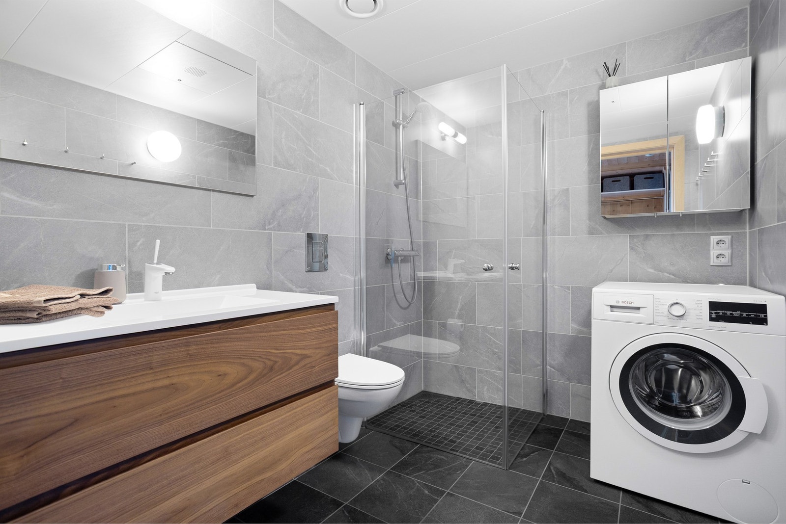 Flislagt baderom med dusjhjørne, vegghengt toalett, innredning med servant og opplegg til vaskemaskin Galleribilde