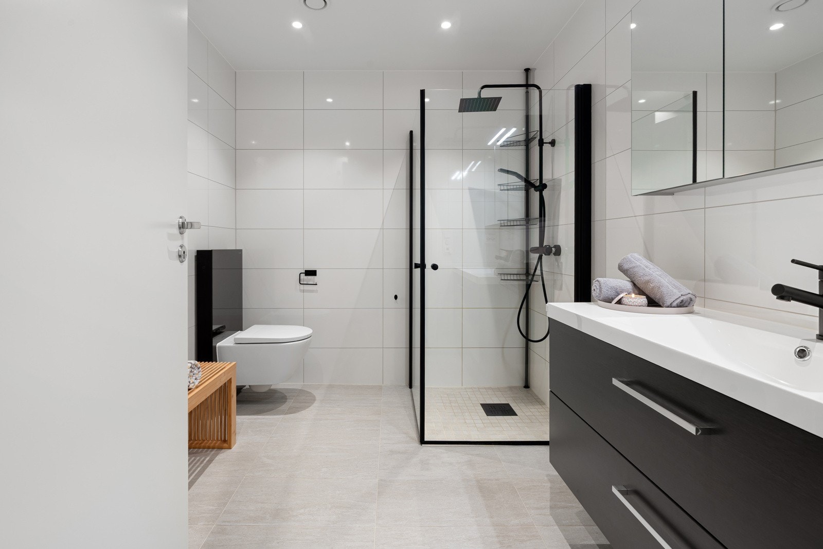 Badet er utstyrt med vegghengt servant, dusjvegger av glass og dusjgarnityr på vegg og vegghengt toalett for et rent og moderne uttrykk.