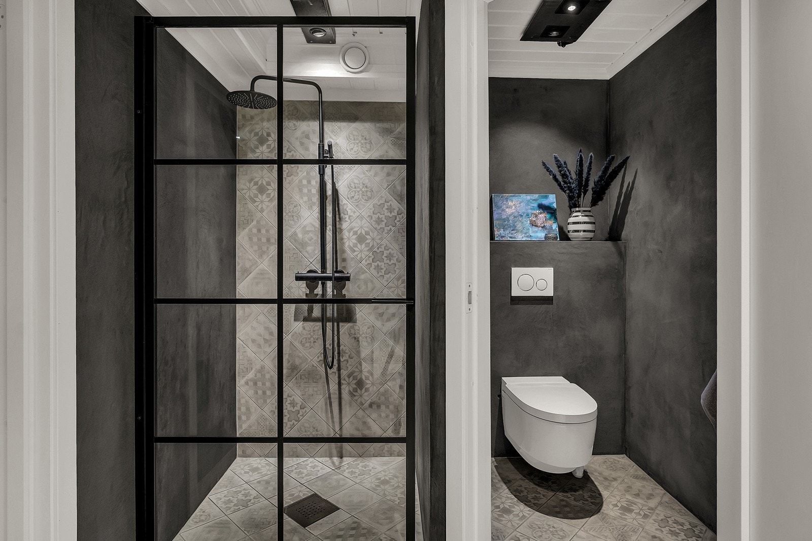 Badet er pent utført med grå mønsterfliser på gulvet og én vegg i dusjsonen, plater med mikrosement i grå finish på øvrige vegger, hvitmalt panelhimling med downlights og dagslys fra et vindu med ruglete glass. Galleribilde