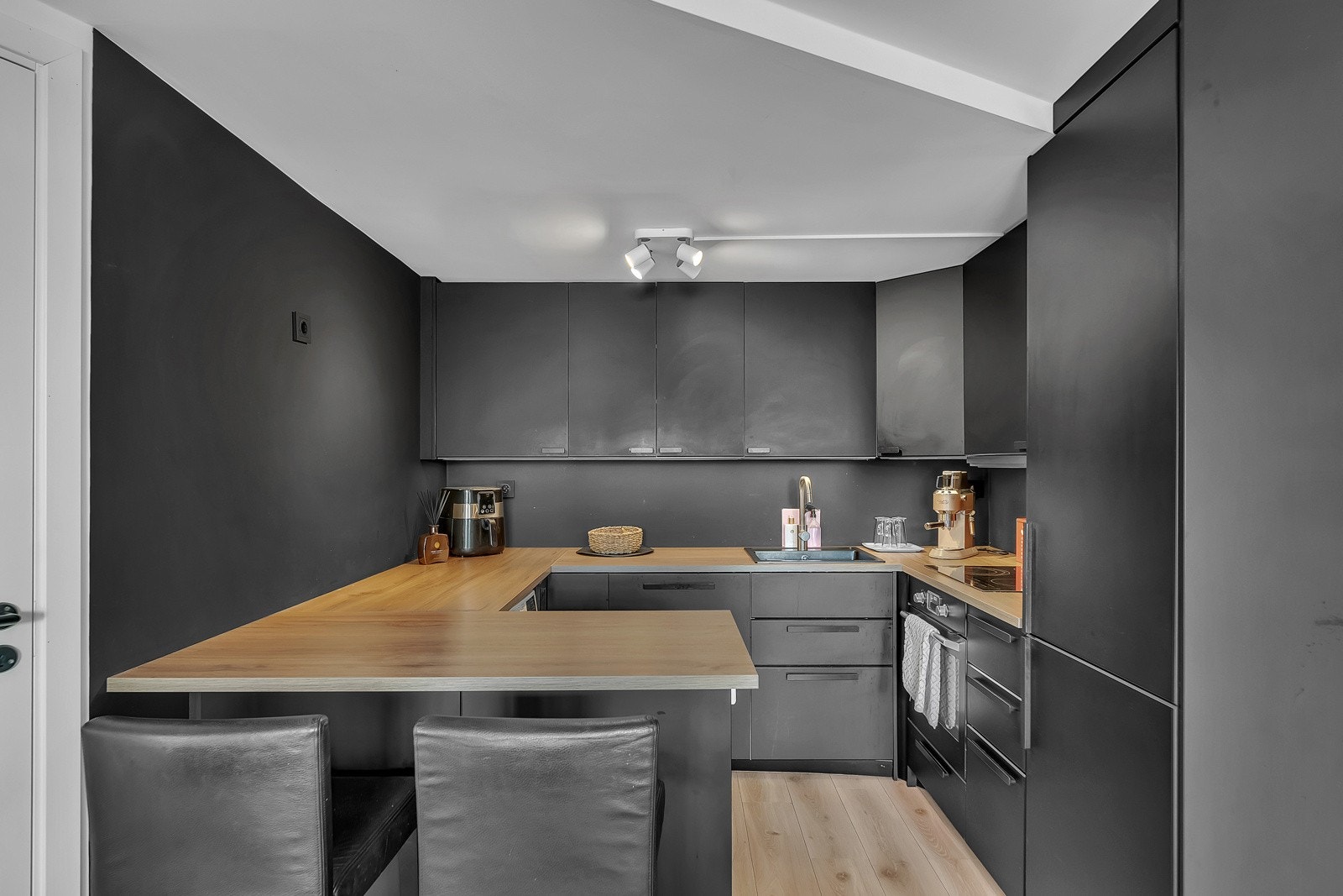 Kjøkkeninnredningen fra 2020 er plassert innerst i rommet og har glatte, mørke fronter i samme nyanse som veggene og en trehvit benkeplate i laminat med nedfelt vaskekum Galleribilde
