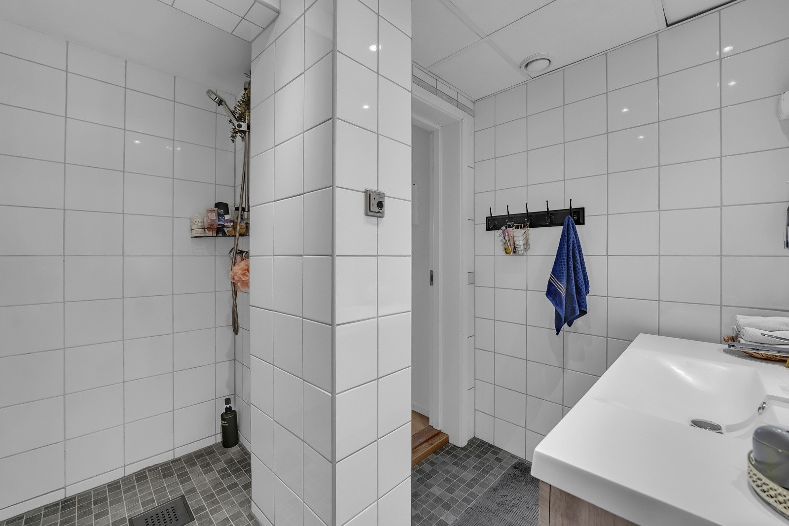 Badet i hybelen har grå mosaikkfliser på gulvet, hvite fliser på veggene og downlights i taket. Innredningen består av en heldekkende servant på skuffeseksjon, et toalett og en dusjnisje Galleribilde