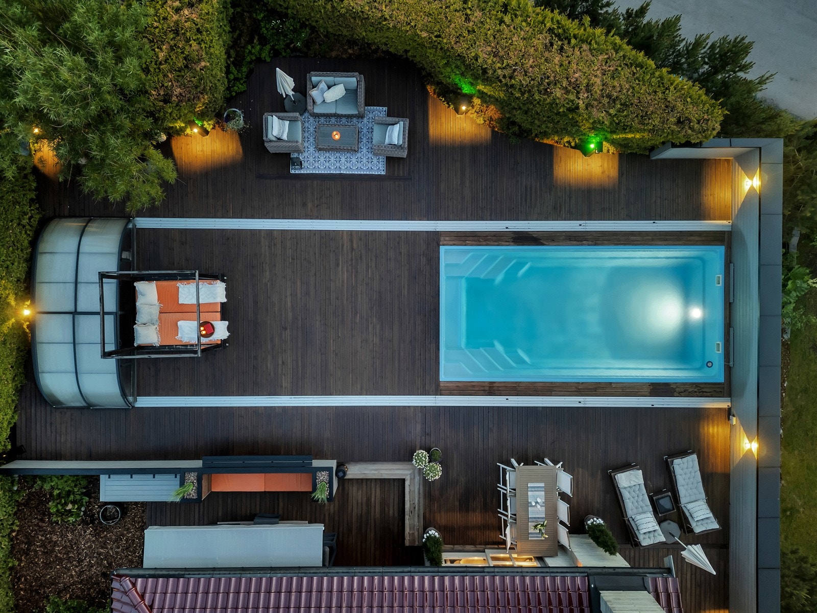 Terrassen rundt om dekke av terrassebord, fin utebelysning, høy levegg langs nordsiden og skjermes ellers av hekken og høye prydbusker Galleribilde