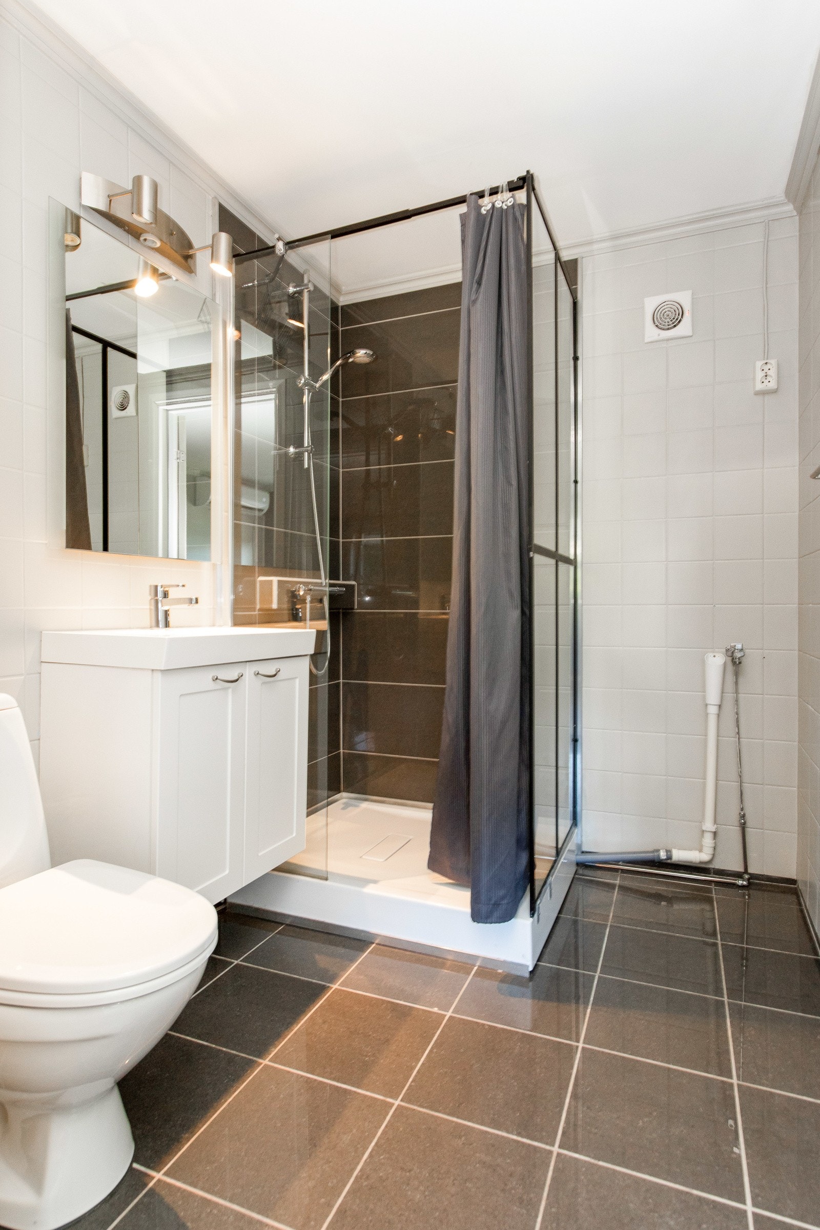 Badet har mørke fliser på gulvet, lyse fliser på veggene, elektrisk avtrekksvifte og opplegg for vaskemaskin Galleribilde