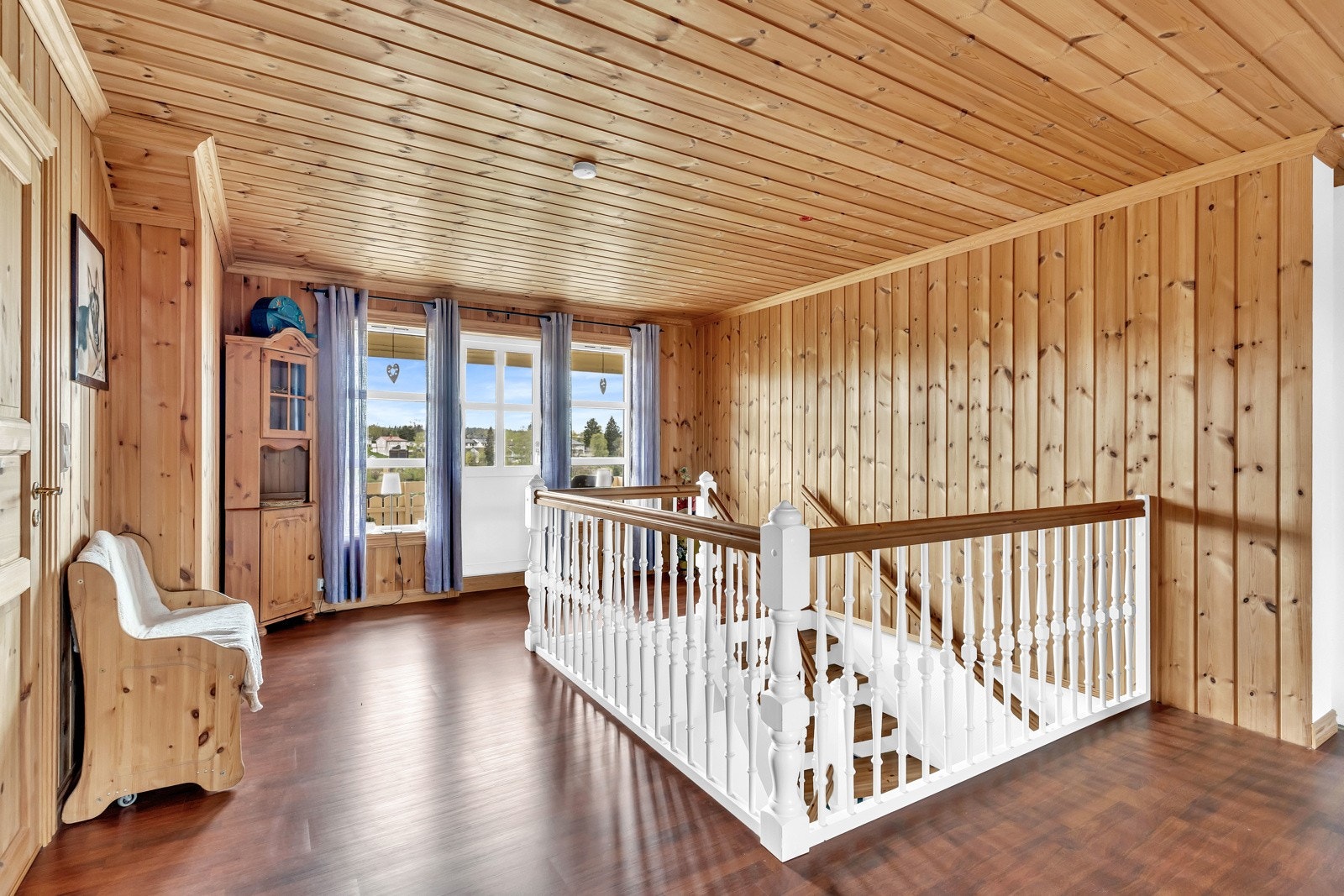 Loftstuen gir et lunt inntrykk med en kombinasjon av laminatgulv, panelte overflater og hvitmalt rekkverk mot trappeløpet. Galleribilde