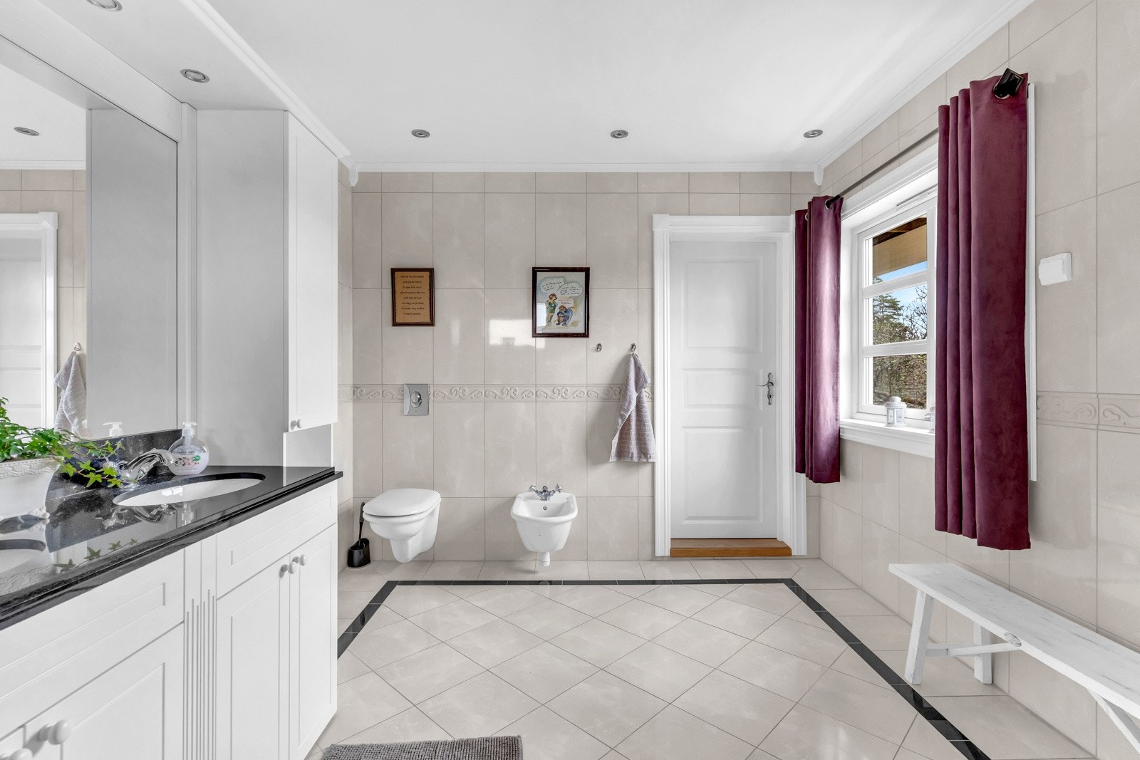 Videre har badet veggmontert toalett og et bidé. Innredningen er hvit med profilerte fronter, to matchende høyskap, sort benkeplate i stein og to servanter. Galleribilde