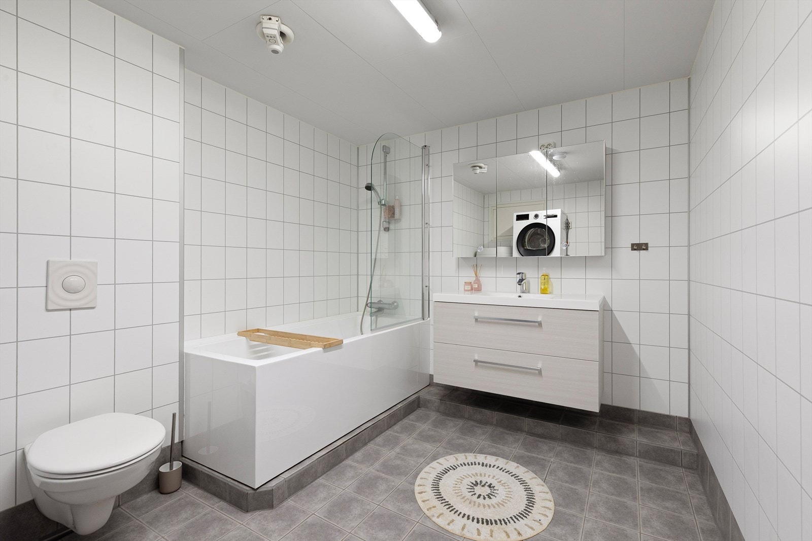 Kombinert vaskerom/bad med fliser på golv og veggar. Rommet er utstyrt med toalett, badekar, servant med innredning og opplegg for vaskemaskin. Galleribilde