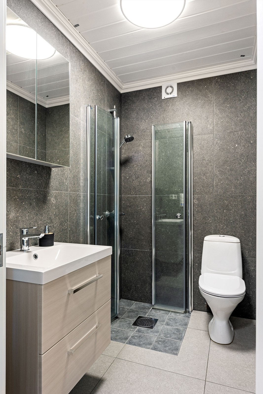 Pent helfliset baderom med varmekabler i gulv. Rommet er utstyrt med servant over skuffeinnredning, speilskap, dusjhjørne med innfellbare dusjvegger og toalett Galleribilde