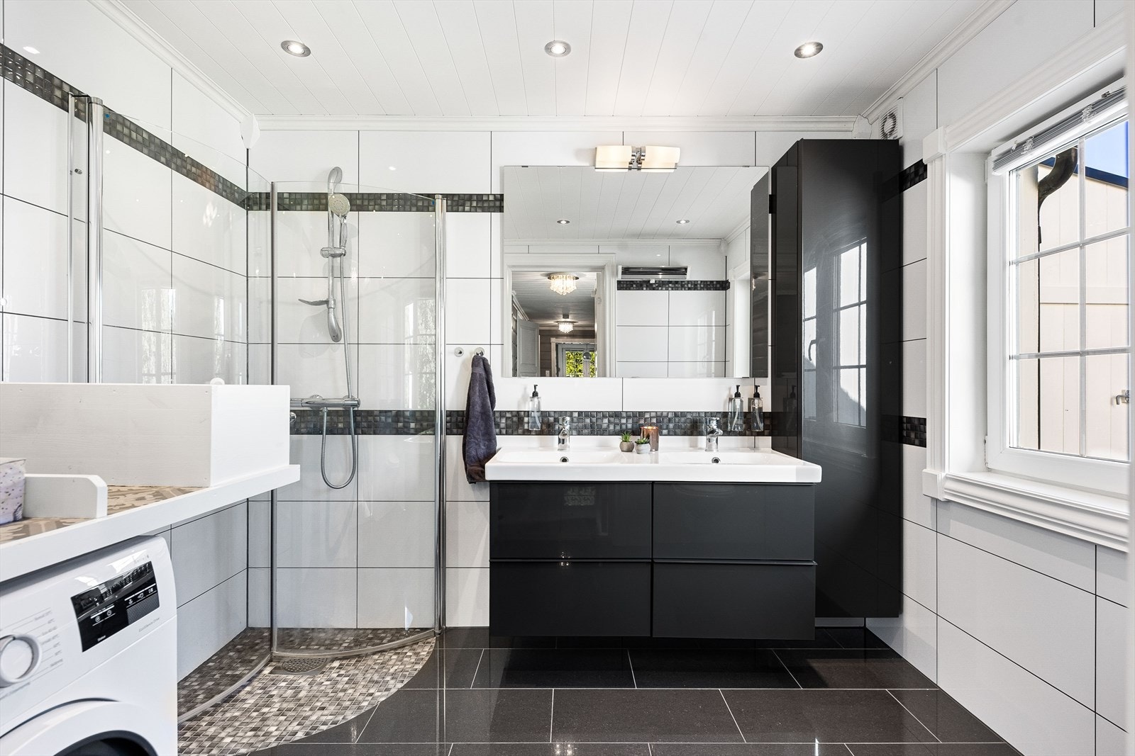 Pent helfliset baderom med varmekabler i gulv. Badet er utstyrt med dobbel servant over skuffeinnredning, speil, vegghengt skap, dusjhjørne med innfellbare dusjvegger, toalett og opplegg for vaskemaskin Galleribilde