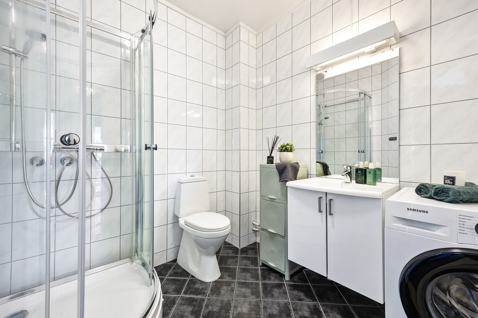 Badet er helfliset med mørke fliser på gulv og hvite på veggene. Rommet er utstyrt med dusjkabinett, toalett og en servant med underskap. Galleribilde