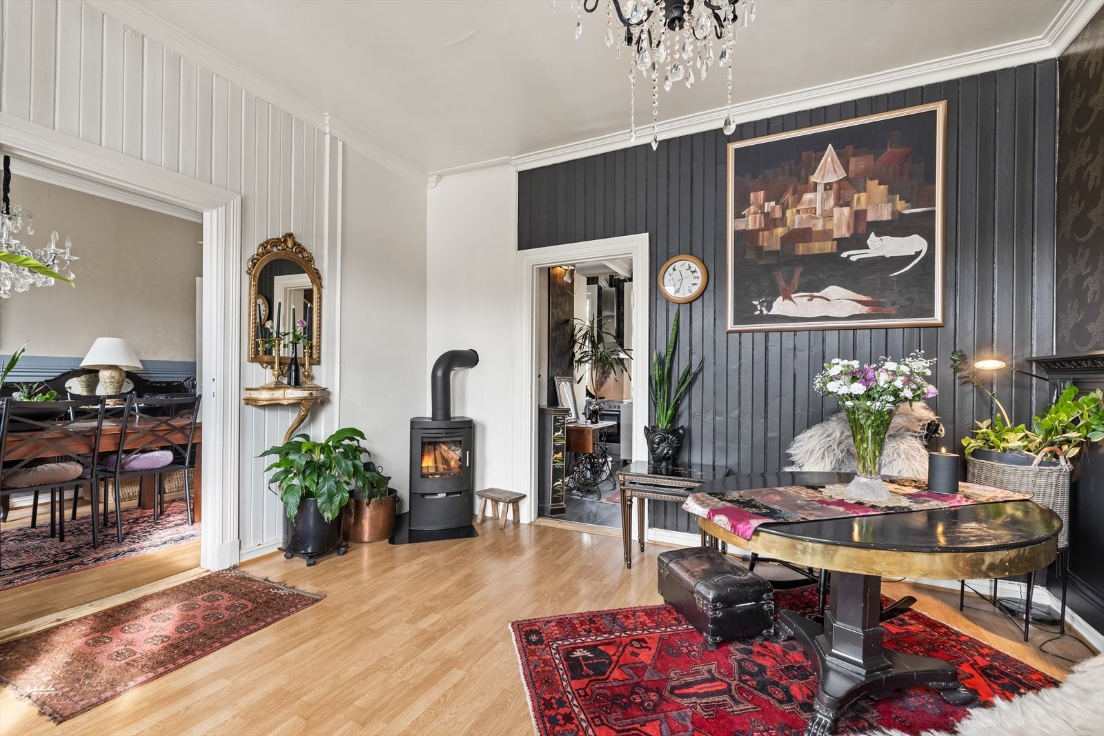 Kontrastvegg med pen tapet og klassisk brystpanel i sort. I stuen er det fine elementer av bredt listverk, dekorert himling, vedovn i hjørnet. Galleribilde