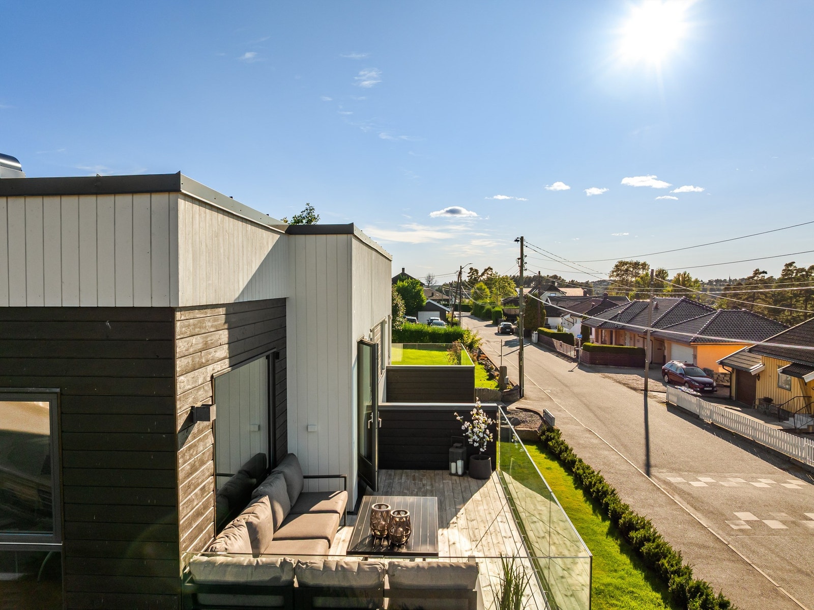 Fasaden med mørk og hvit trekledning rammer inn en romslig terrasse med åpen utsikt mot rolig villastrøk. Galleribilde