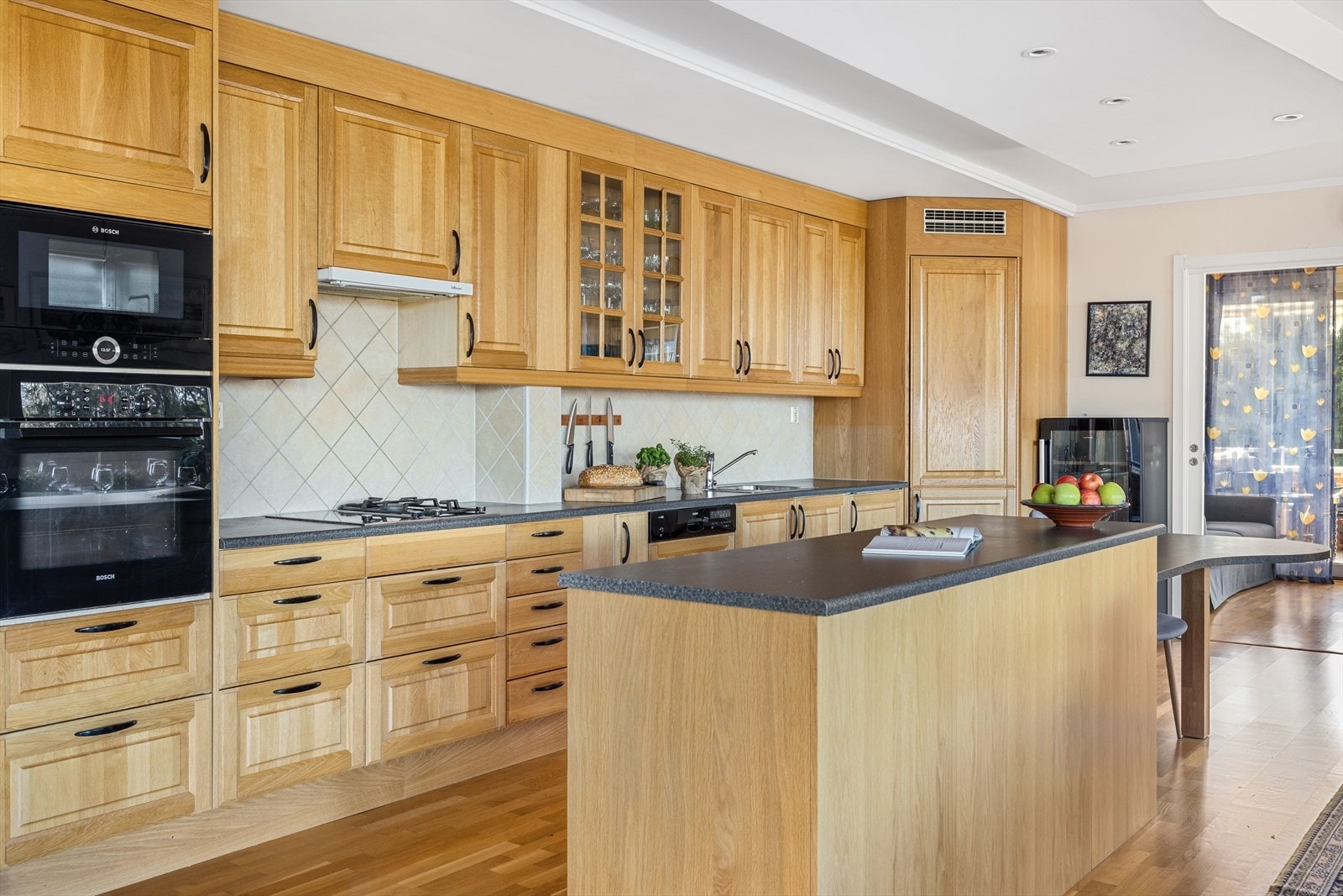 Dette er et delikat og velholdt kjøkken med innredning fra Norema i heltre eik. En praktisk kjøkkenøy deler av kjøkkenet ut mot spiseplassen, samtidig som det gir både ekstra lageringsplass og en god arbeidsbenk. Galleribilde